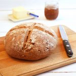 Walnutty Wholemeal Bread on a chopping board with knife with jam & butter in background.