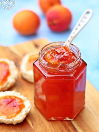 Jar of Peach & Apricot Jam with a spoon and scones on a wooden board surrounded by peaches, nectarines and apricots.