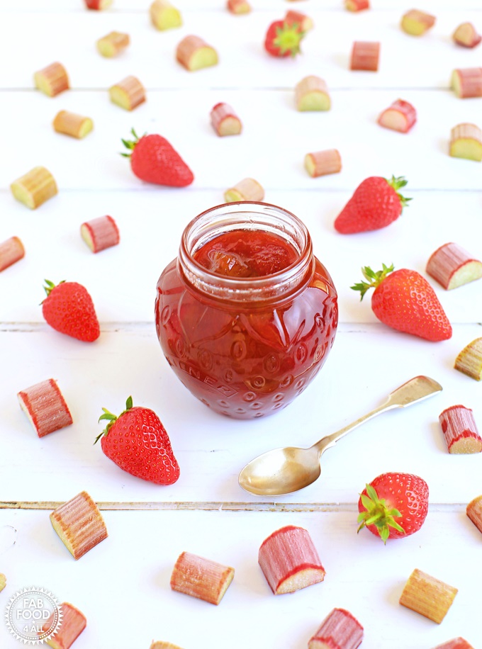 Pot of Rhubarb & Strawberry Jam with teaspoon surrounded by strawberries & slices of rhubarb.