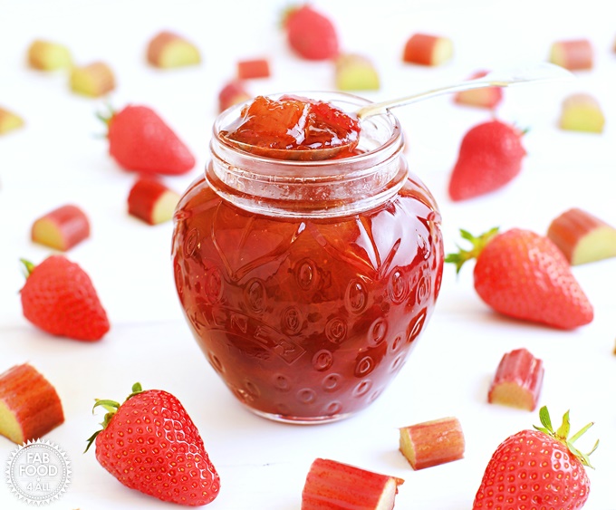 Pot of Rhubarb & Strawberry Jam with teaspoon surrounded by strawberries & slices of rhubarb.