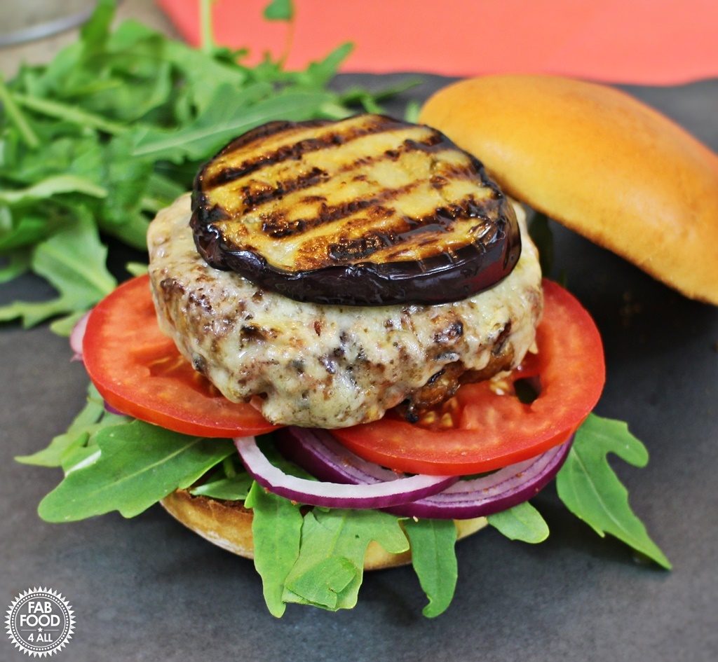 Welsh Lamb Moussaka Burger on a bed of rocket, red onion and sliced tomato with melted cheese and grilled aubergine on top with brioche bun top tilted to one side.