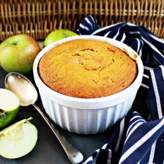 Eve's Pudding in a white dish surrounded by Bramley apples, a tea towel & serving spoon.
