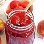 Jar of Strawberry & Peach Jam with teaspoon & fruits in background.
