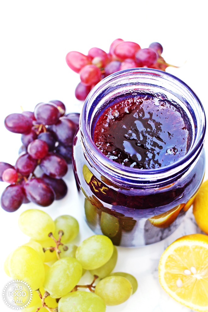 Opened jar of Easy Grape Jam surrounded by a variety of seedless grapes.