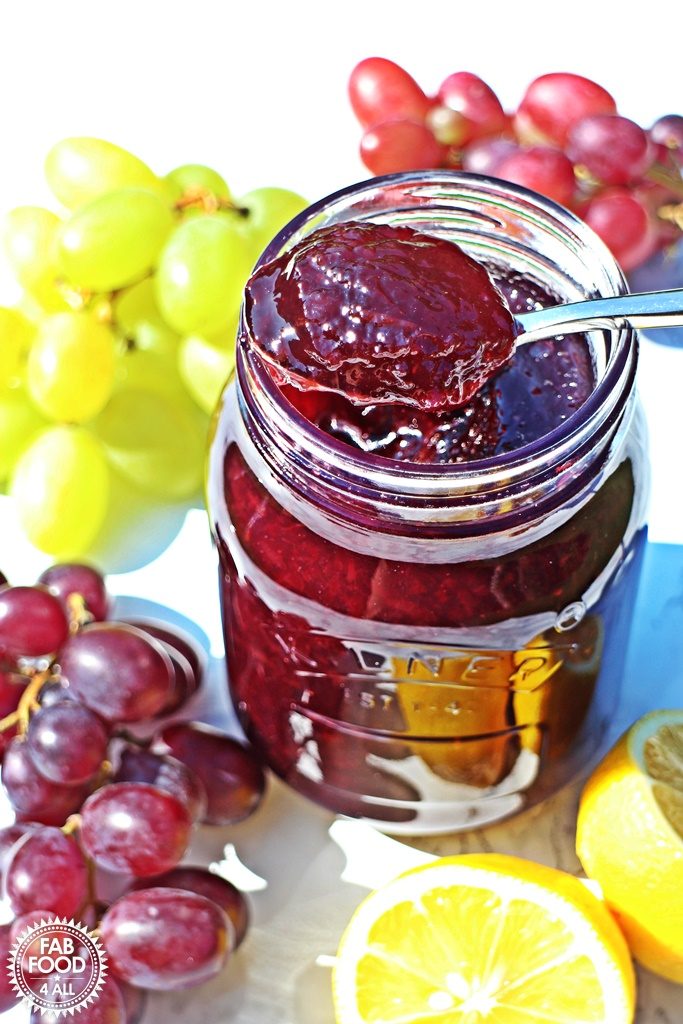 Opened jar of Easy Grape Jam with teaspoon of jam resting on top surrounded by grapes.