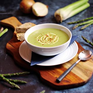 Cream of Asparagus & Celery Soup on a wooden board surrounded by asparagus, celery and onion.