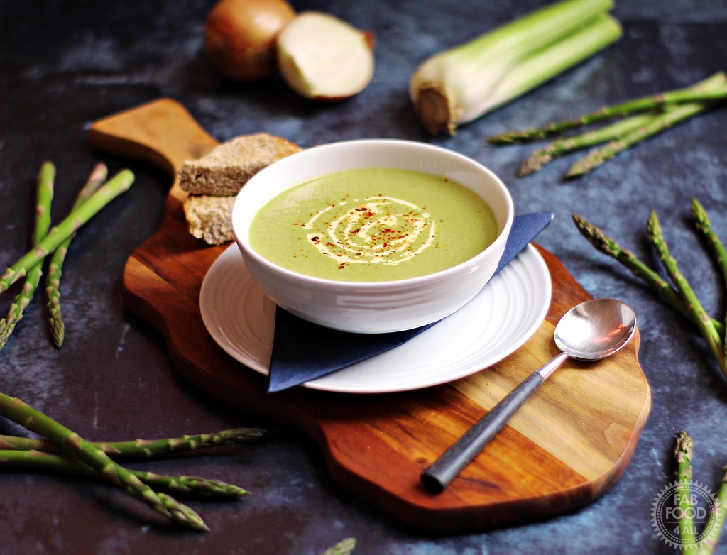 Cream of Asparagus & Celery Soup on a wooden board surrounded by asparagus, celery and onion.