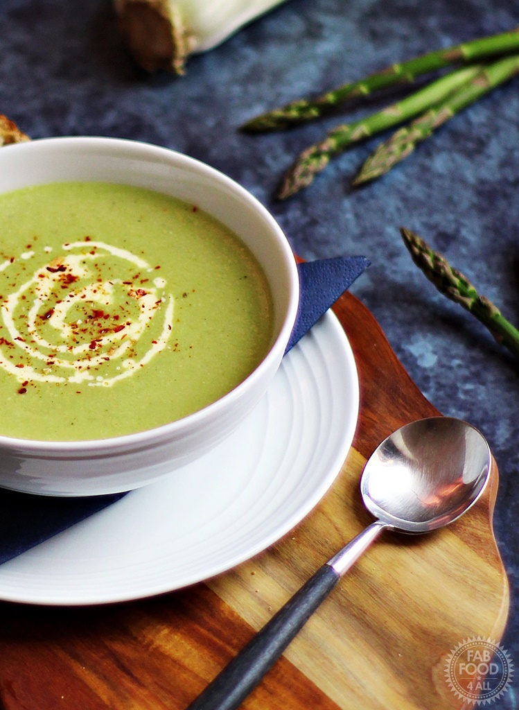 Cream of Asparagus & Celery Soup on a wooden board surrounded by asparagus, celery and onion.