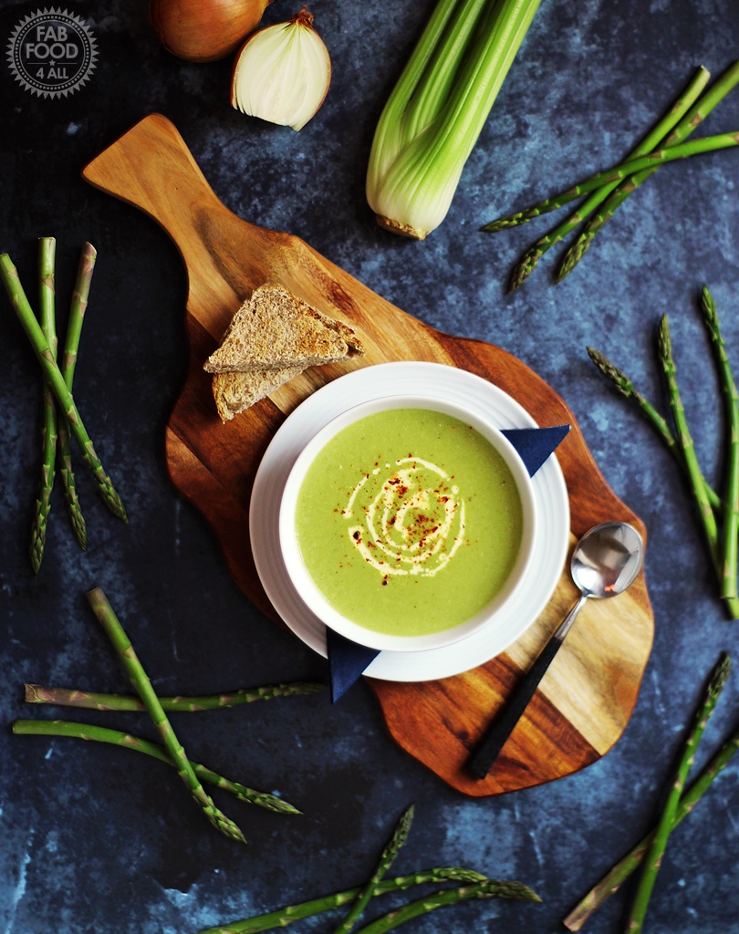 Aerial view of Cream of Asparagus & Celery Soup on a wooden board surrounded by asparagus, celery and onion.