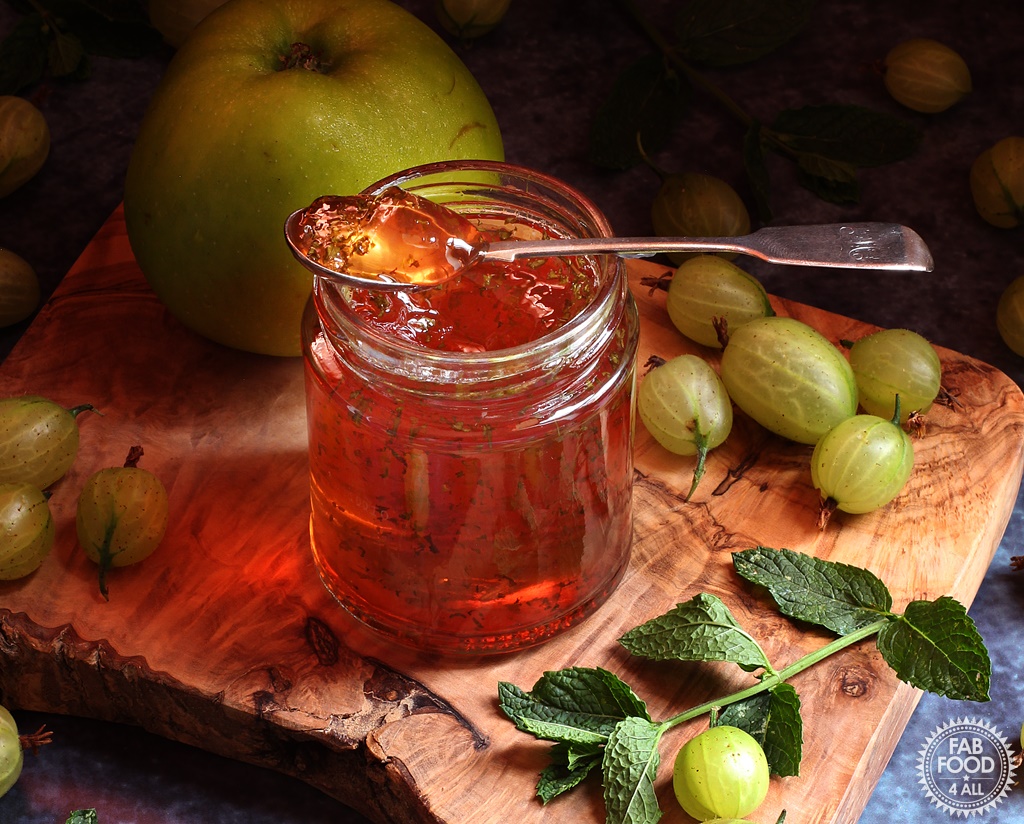Gooseberry, Apple & Mint Jelly on a board surrounded by gooseberries, Bramley apple, and mint leaves.