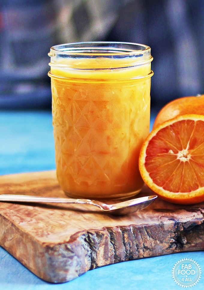 Granny's Quick Blood Orange Curd in a glass jar on wooden board with spoon and blood oranges.