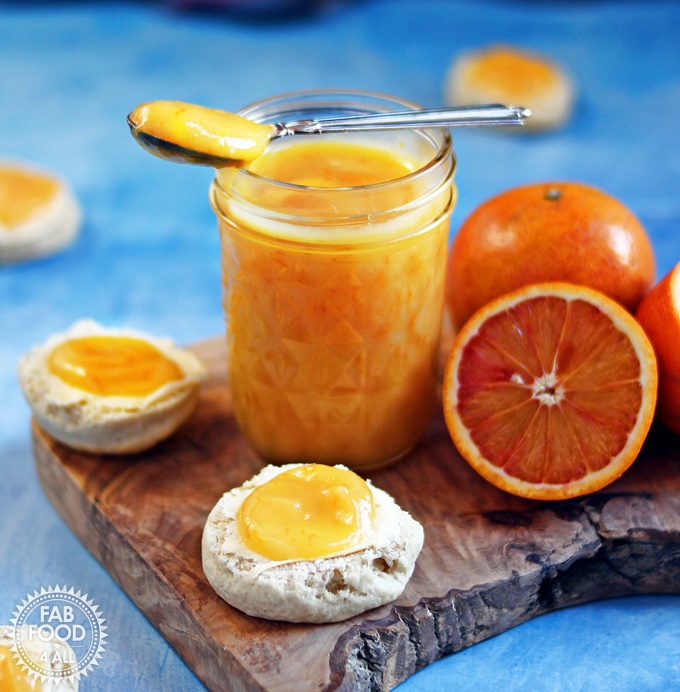 Granny's Quick Blood Orange Curd in a glass jar on wooden board with spoon and blood oranges with scones scattered around.