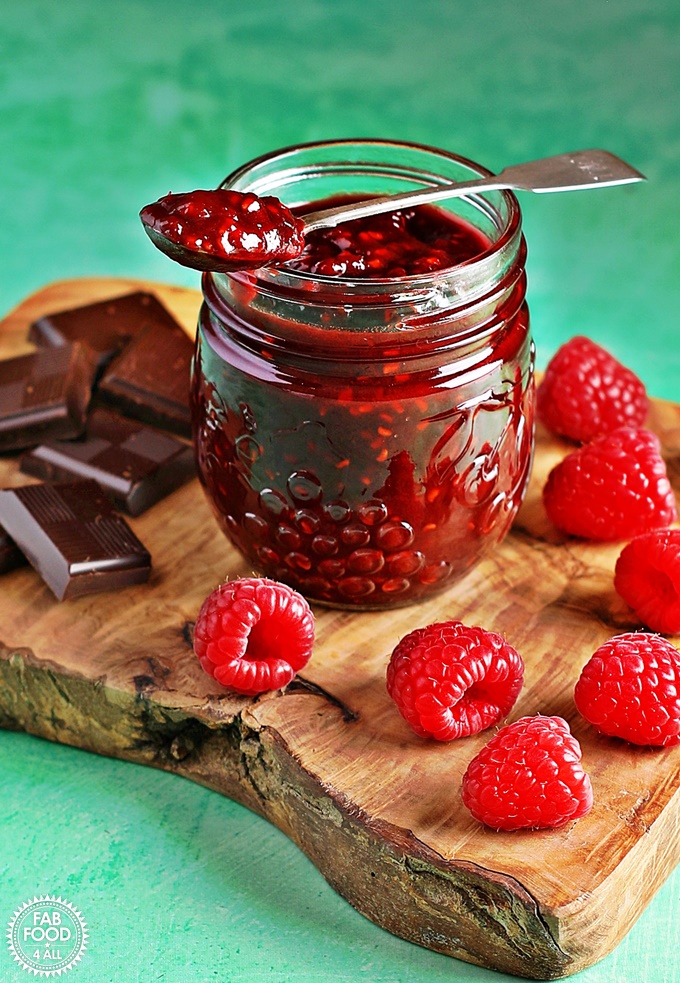 Jar of Raspberry & Chocolate Jam on a wooden board with dark chocolate & raspberries.
