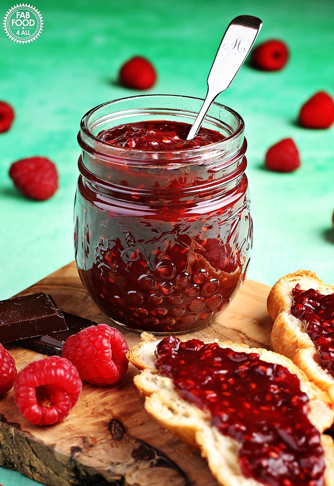 Jar of Raspberry & Chocolate Jam on a wooden board with dark chocolate & raspberries.