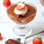 Foolproof Rich Chocolate Mousse with piped whipped cream & strawberry decoration in a sundae glass with spoonful of mousse on side.