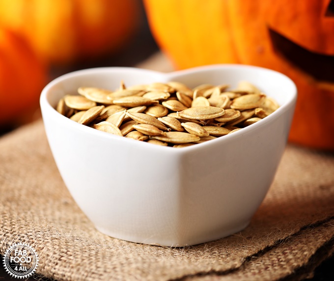 Close up or Roasted Pumpkin Seeds in a dish with Pumpkins & jack-o-lantern in the background.