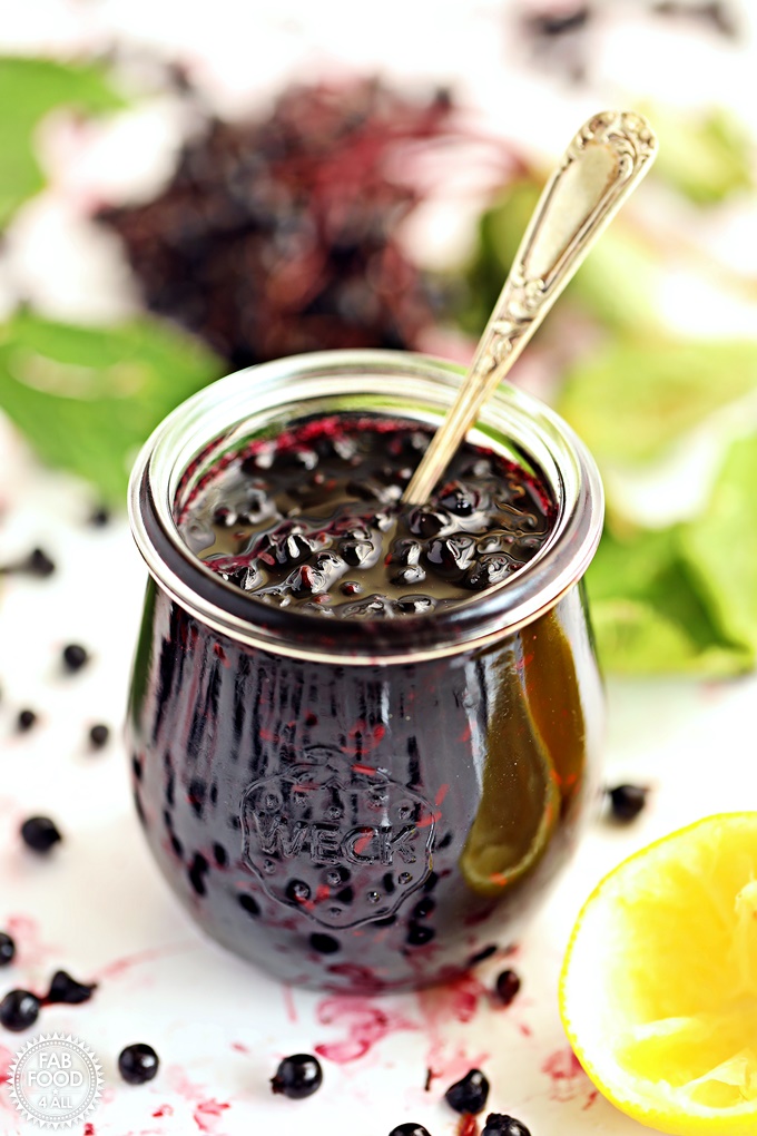 Elderberry Jam in jar with teaspoon submerged, surrounded by blueberries & lemon rinds.