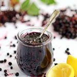 Blueberry Jam in jar with teaspoon submerged, surrounded by blueberries & lemon rinds.