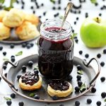 Sloe & Apple Jam in a jar on plate with spoon in jar, scones and apple in background.