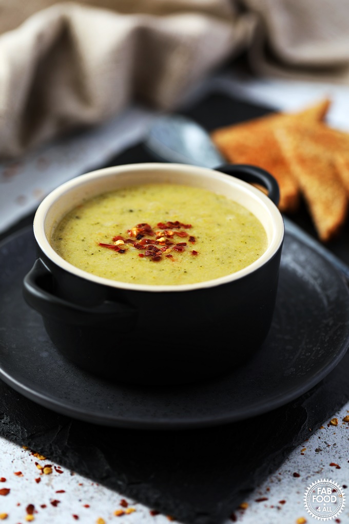 Broccoli & Cheddar Soup in a bowl on a plate with spoon & toast on a slate.