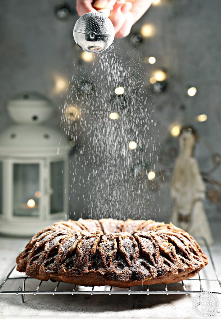 Christmas Bundt Cake being dusted with icing sugar (with Christmas lights, angel decoration and white lantern in background).