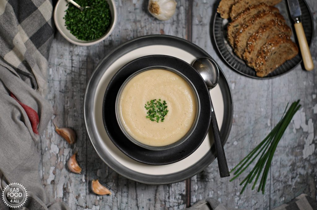 Overhead shot of Easy Roasted Celeriac Soup in a bowl with sliced bread, scattered garlic and chive garnish in a bowl.