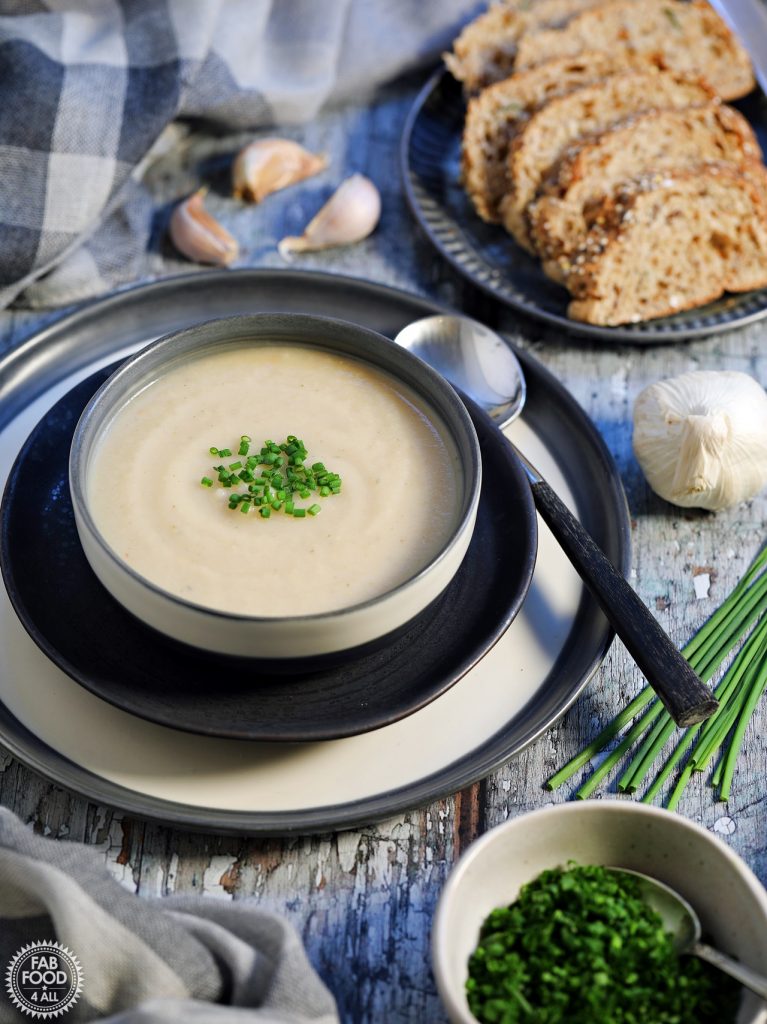 Overhead shot of Easy Roasted Celeriac Soup in a bowl with sliced bread, scattered garlic and chive garnish in a bowl.
