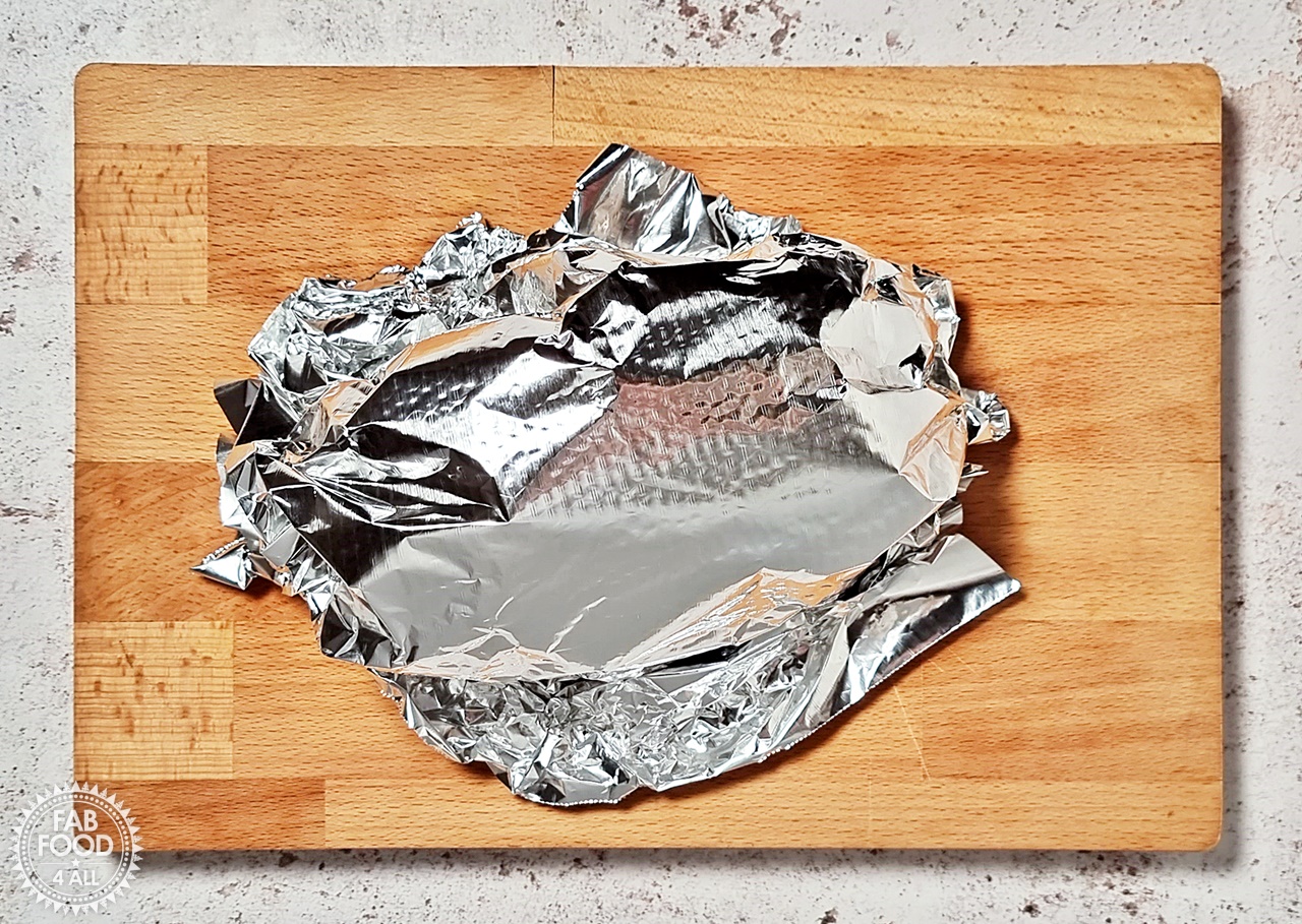 Beef brisket on a wooden board covered in foil to rest.