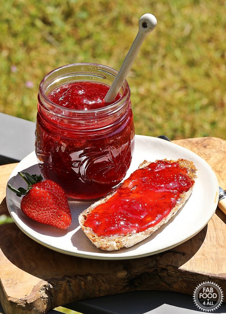 Strawberry & Gin Jam jar on a plate with bread & jam & a strawberry with lawn in background.