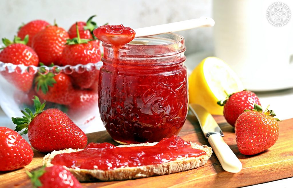 Strawberry & Gin Jam jar with spoonful of jam resting on top on a board with bread & jam (bowl of strawberries, cut lemon & mug in background).