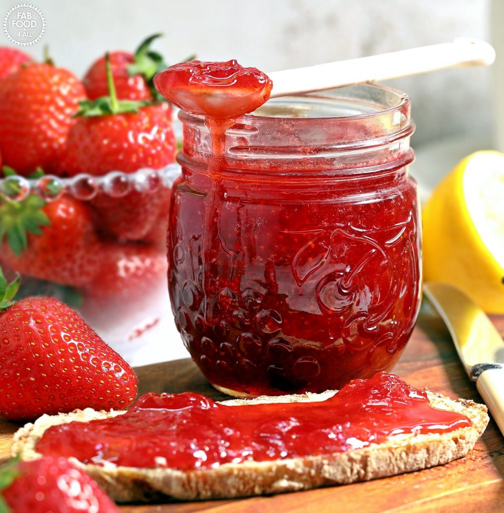 Strawberry & Gin Jam jar with spoonful of jam resting on top on a board with bread & jam (bowl of strawberries and cut lemon in background).