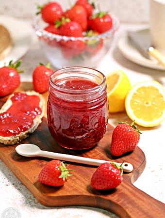 Jar of Strawberry & Gin Jam on a board with bread and jam, strawberries and cut lemon in background..
