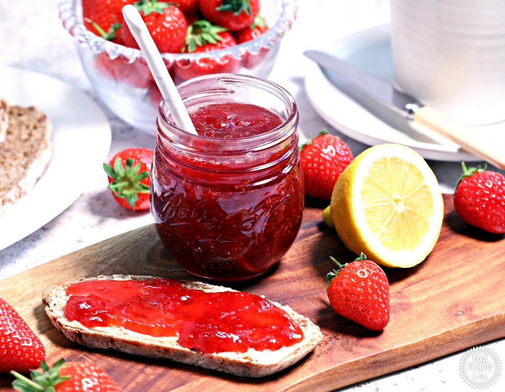 Strawberry & Gin Jam on a wooden board with bread, strawberries and cut lemon.