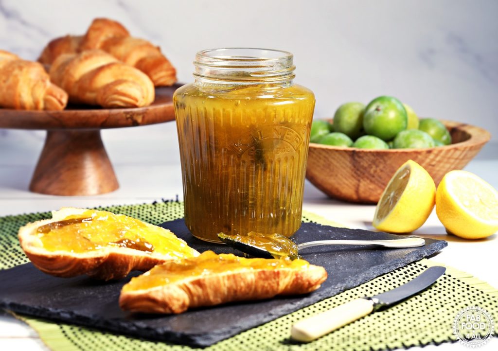 Jar of Easy Greengage Jam on a slate with buttered croissant spread with jam. Bowl of greengages and cake stand of croissants in background.