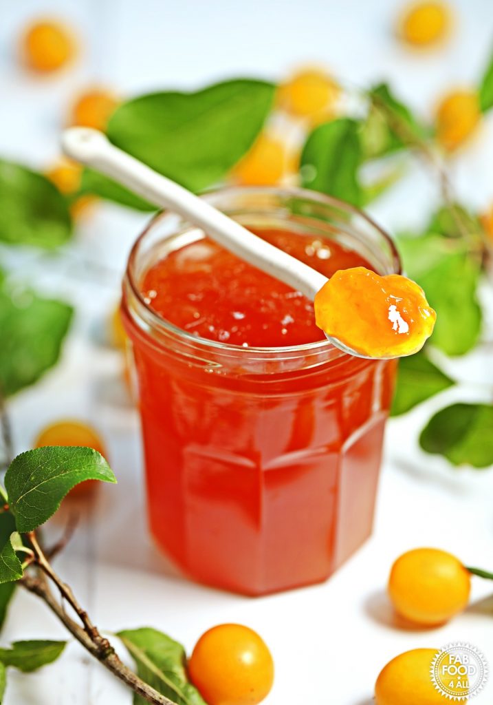 Jar of Mirabelle Plum Jam with spoonful resting on top surrounded by Mirabelles and foliage.