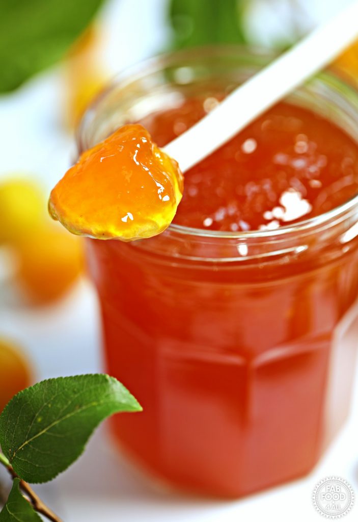 Close up of jar of Mirabelle Plum Jam with spoonful resting on top surrounded by Mirabelles and foliage.