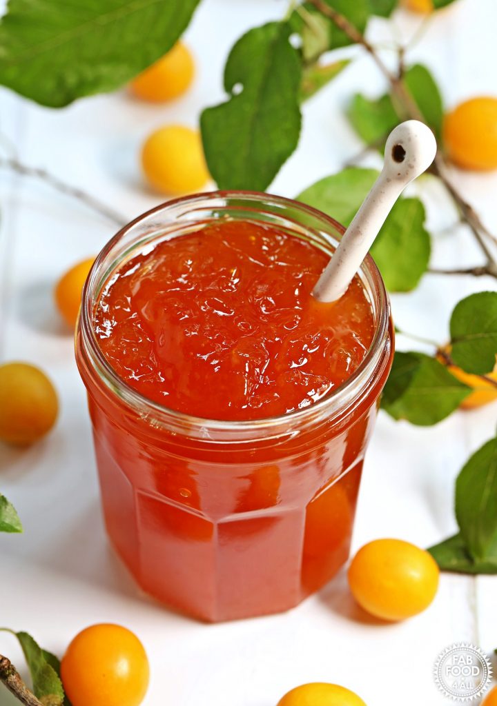 Close up of jar of Mirabelle Plum Jam with teaspoon stood up inside surrounded by Mirabelles and foliage.