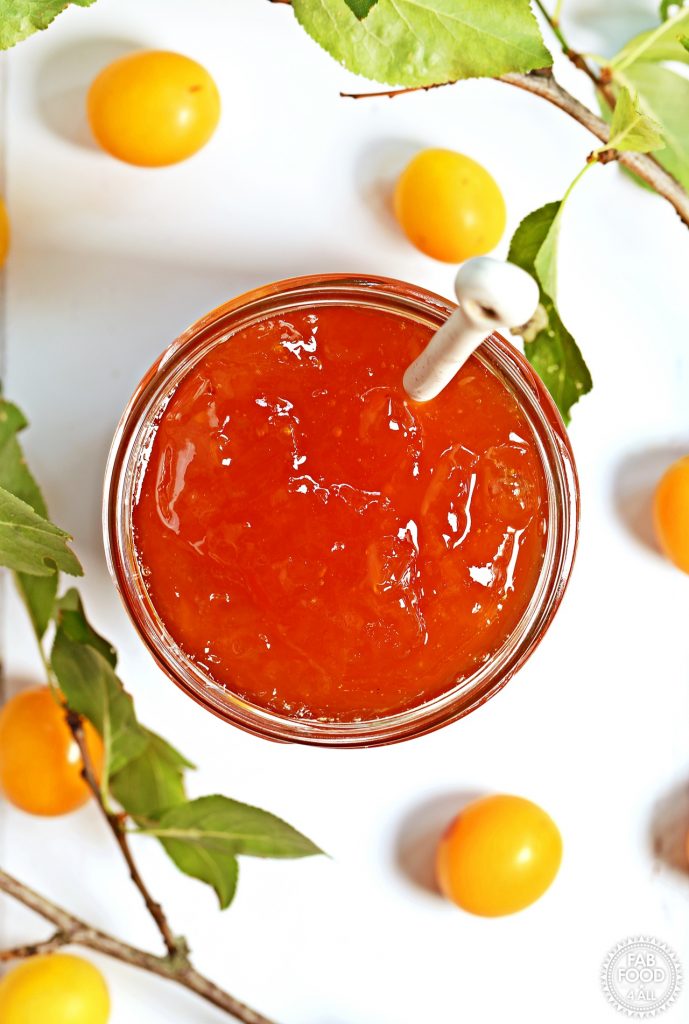 Overhead shot of Mirabelle Plum Jam in a jar with teaspoon surrounded by Mirabelle plums and foliage.