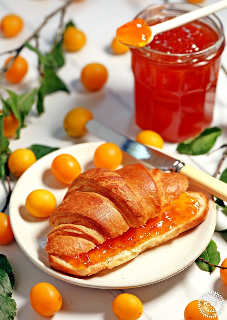 Mirabelle Plum Jam spread on a croissant with jar and Mirabelles in background.