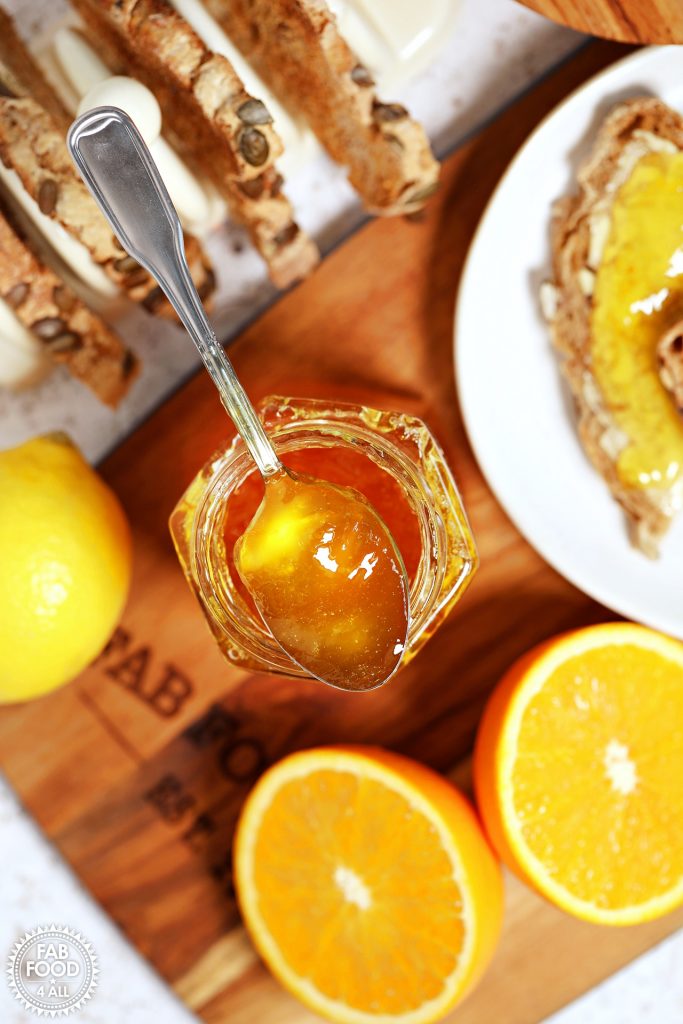 Overhead vertical shot of teaspoonful of Shredless Orange Marmalade resting on top of hexagonal jar surrounded by toast, lemon and cut oranges.