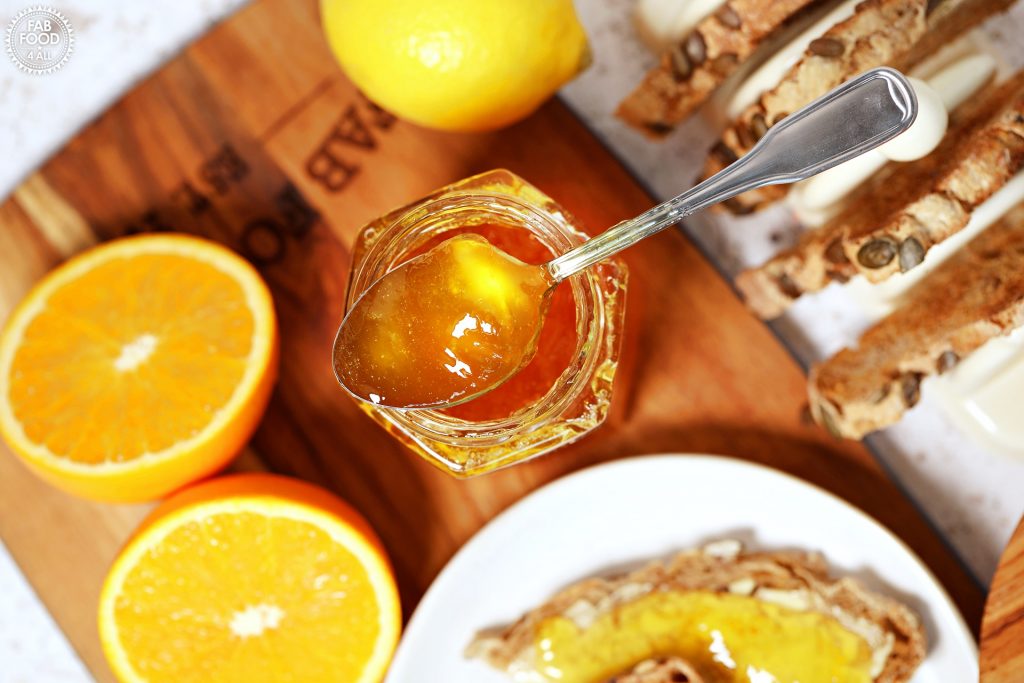 Overhead shot of teaspoonful of Shredless Orange Marmalade resting on top of hexagonal jar surrounded by toast, lemon and cut oranges.