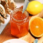 Jar of Simple Shredless Orange Marmalade with a teaspoon in it on a board surrounded by lemon, cut orange and toast.