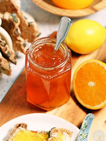 Jar of Simple Shredless Orange Marmalade with a teaspoon in it on a board surrounded by lemon, cut orange and toast.