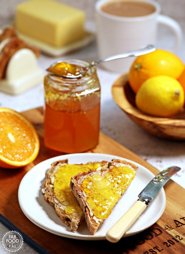 Simple Shredless Orange Marmalade on toast on a plate with jar, bowl of fruit, butter, cup of tea and toast rack in background.