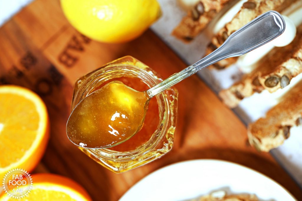Close up overhead shot of teaspoonful of Shredless Orange Marmalade resting on top of hexagonal jar surrounded by toast, lemon and cut oranges.