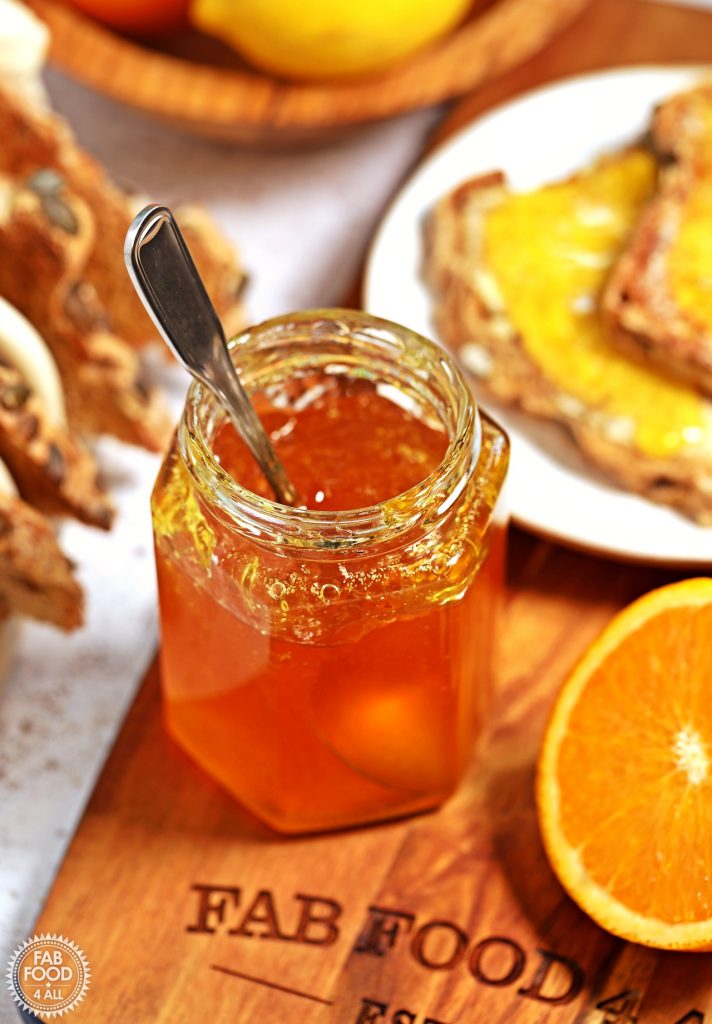 3/4 angle shot of jar of Shredless Orange Marmalade with spoon in it on a wooden board surrounded by toast, cut orange and fruit bowl.
