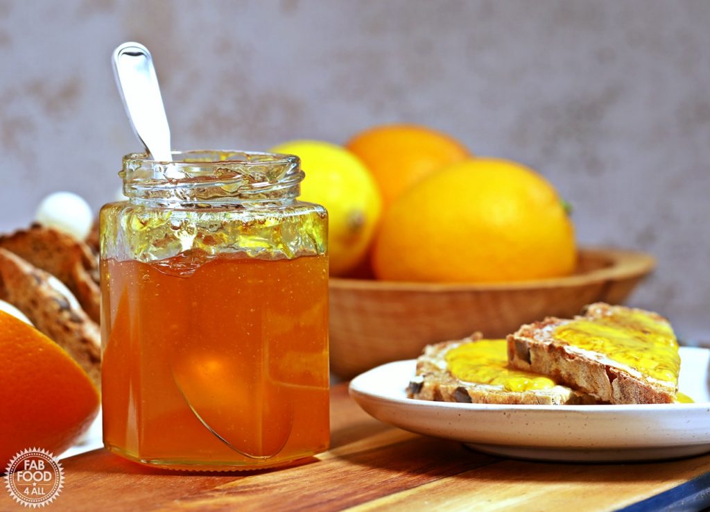Side on shot of jar of Simple Shredless Orange Marmalade with a teaspoon in it on a board with plate of toast and bowl of citrus fruits in background.