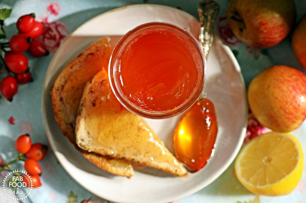 Overhead shot of jar of Rosehip & Apple Jam on a plate with toast and teaspoon surrounded by rosehips, apple and lemon.
