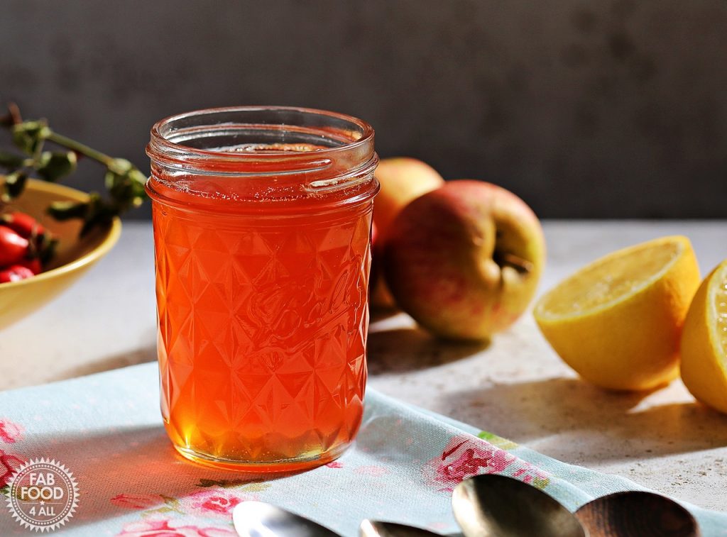 Jar of Rosehip & Apple Jelly with apples, rosehips and lemon in background.