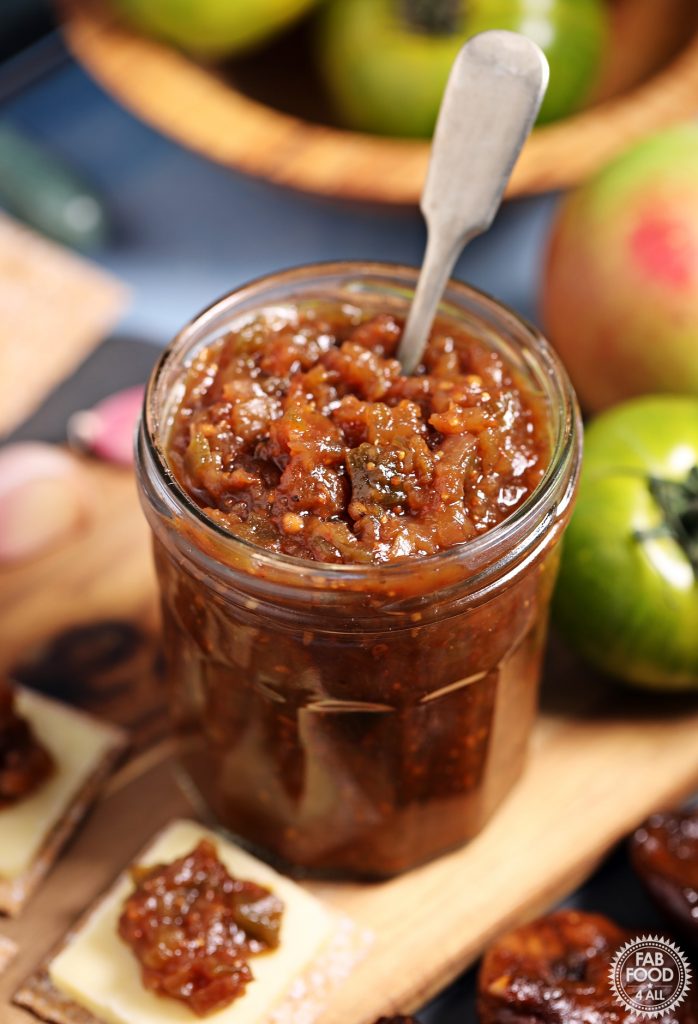 Easy Green Tomato Chutney in a jar with teaspoon inserted, on a board with cheese & crackers.
